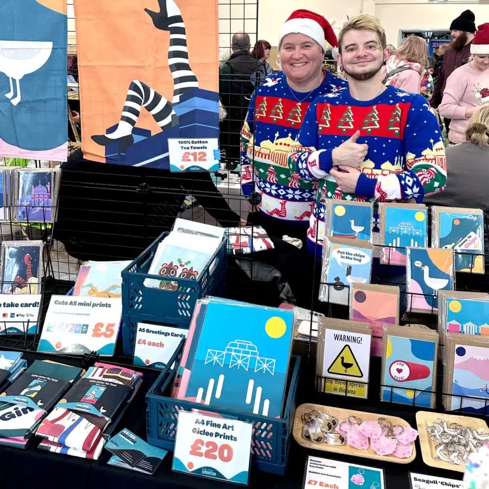 Two independent makers in Christmas jumpers smiling behind a colourful stall of prints, cards, tea towels and gifts at a Shoreham Christmas makers fair.