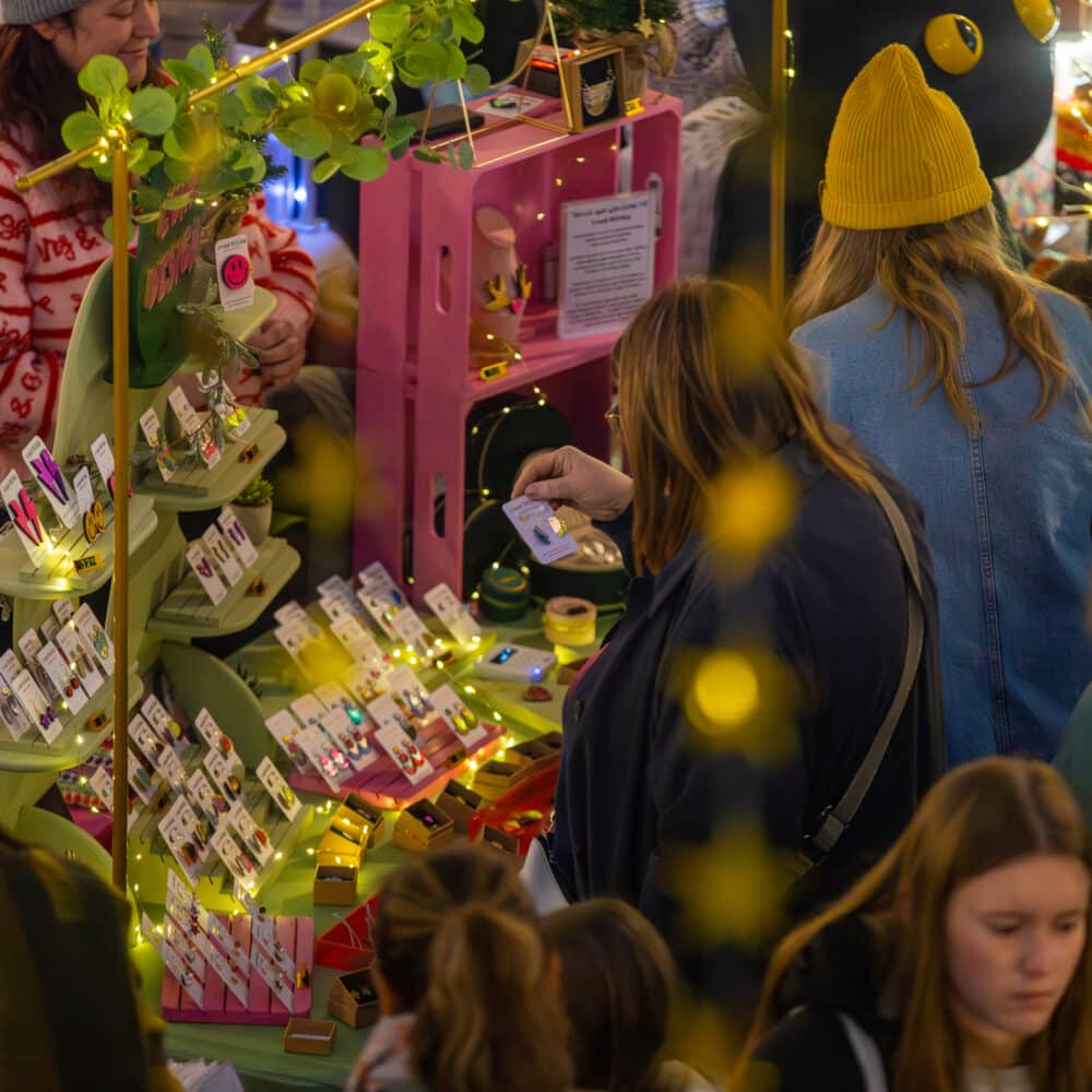 Shoppers browsing a colourful independent jewellery and accessories stall decorated with fairy lights at a busy Paper Daisy Events makers fair.