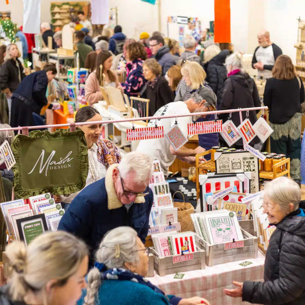 Busy indoor makers fair at The Shoreham Centre with visitors browsing colourful stalls selling art, prints, cards and handmade gifts.