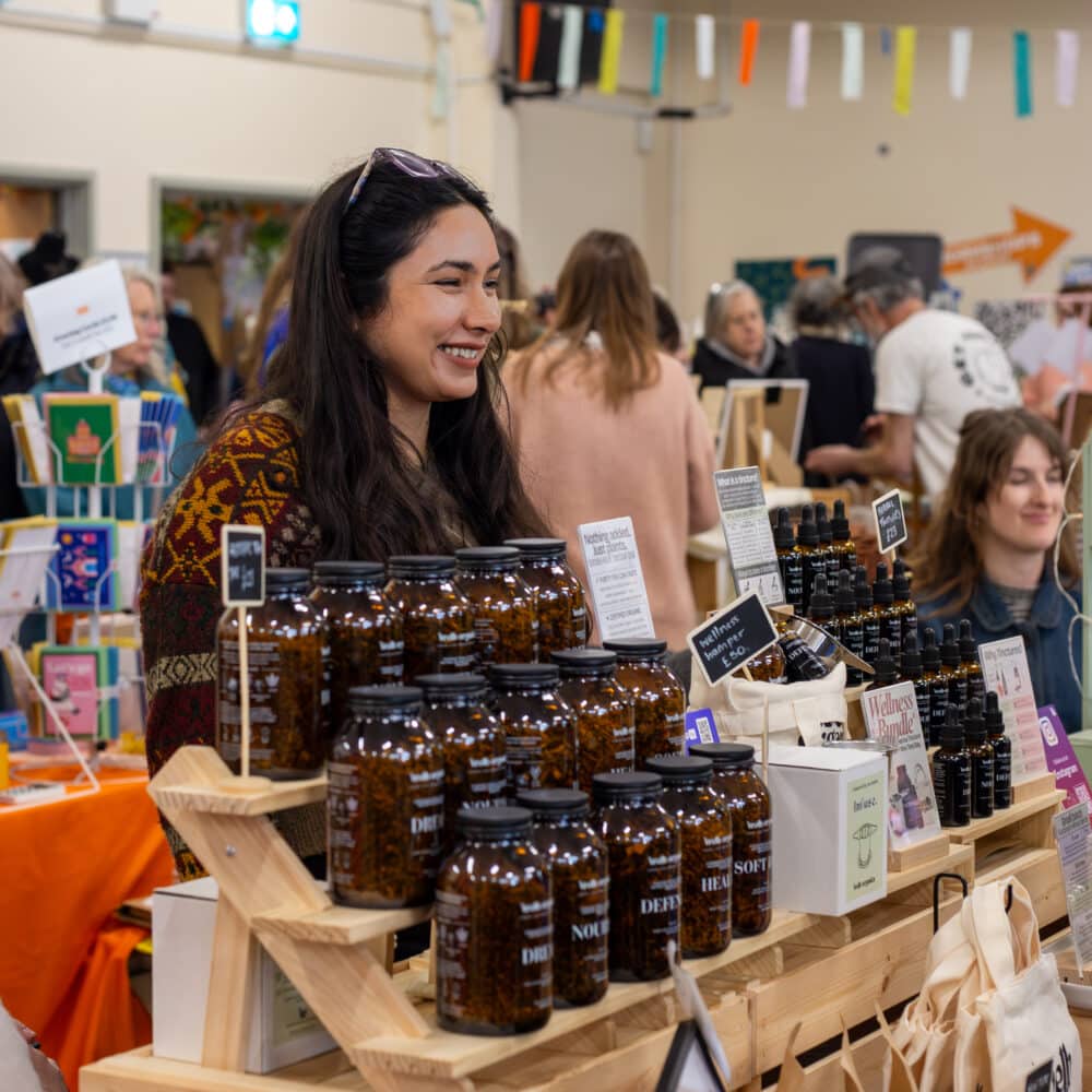 Smiling independent maker behind a stall of handmade wellness and self-care products at a Paper Daisy Events makers fair in Shoreham.
