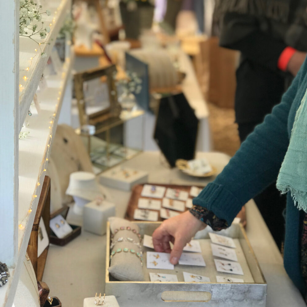 A shopper dressed in green and blue reaches out to pick up jewellery from a market stall. The stall is displaying lots of handmade jewellery and is decorated with twinkling fairy lights, white flowers and a linen table cloth..