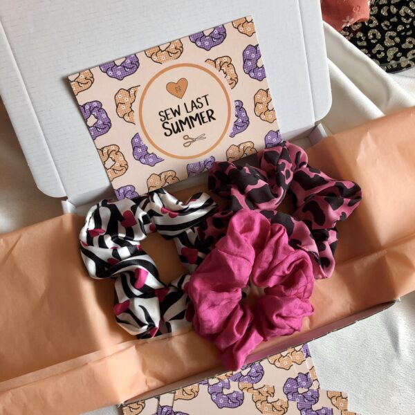 Three scrunchies on peach tissue paper. The scrunchies are: zebra print satin with pink hearts, pink snakeskin effect, and pink and black leopard print.