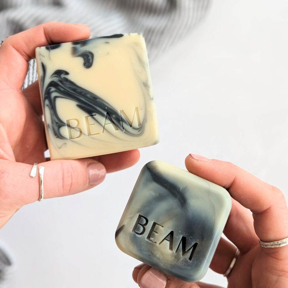 Two white natural soaps with black swirls, one large and one small. Both soaps are square and imprinted with "BEAM". The soaps are held by a person's hands against a light coloured background.