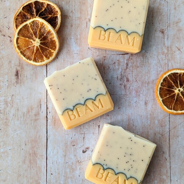 Top down photograph of three square soap bars on a natural wood look background. The soaps are orange on the bottom third and cream on the top, speckled with poppy seeds. The soap contains a black scalloped line. There are three dried orange slices next to the soaps.