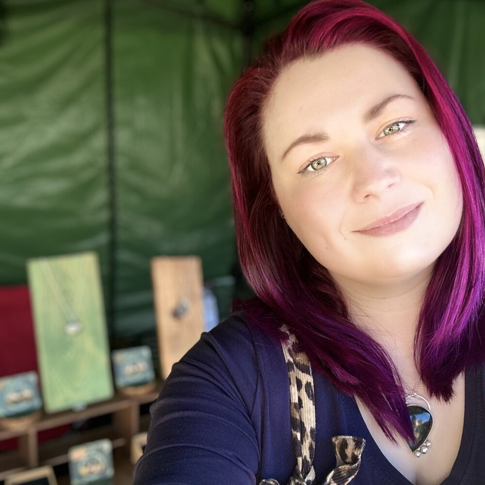 Fran Barnett Jewellery. A woman with pink hair smiles in front of a market stand selling jewellery