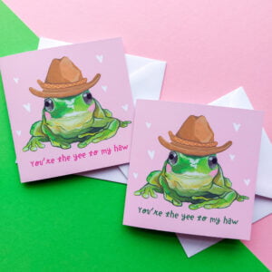 Pink square greeting card with green, blushing tree frog wearing a cowboy hat. Text below reading 'You're the yee to my haw'