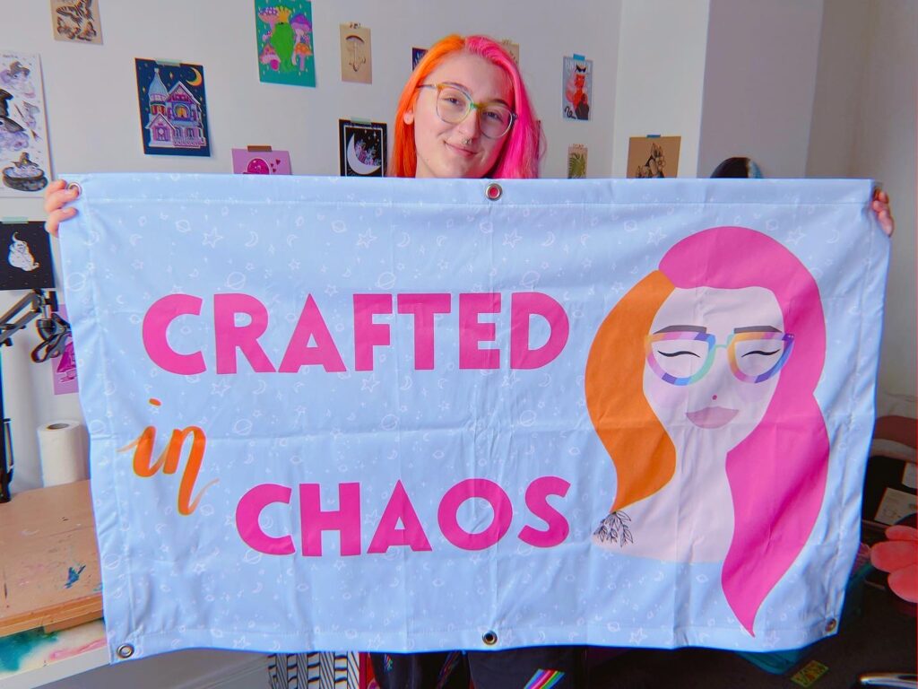 Crafted in Chaos - Meg a white female with pink and orange hair and glasses smiles whilst holding up a large light blue banner with the words Crafted in Chaos in bright orange and pink bold letters, and a digitalised image of Meg in cartoon style - What sort of banner should I use on my stall? 4 Top Tips