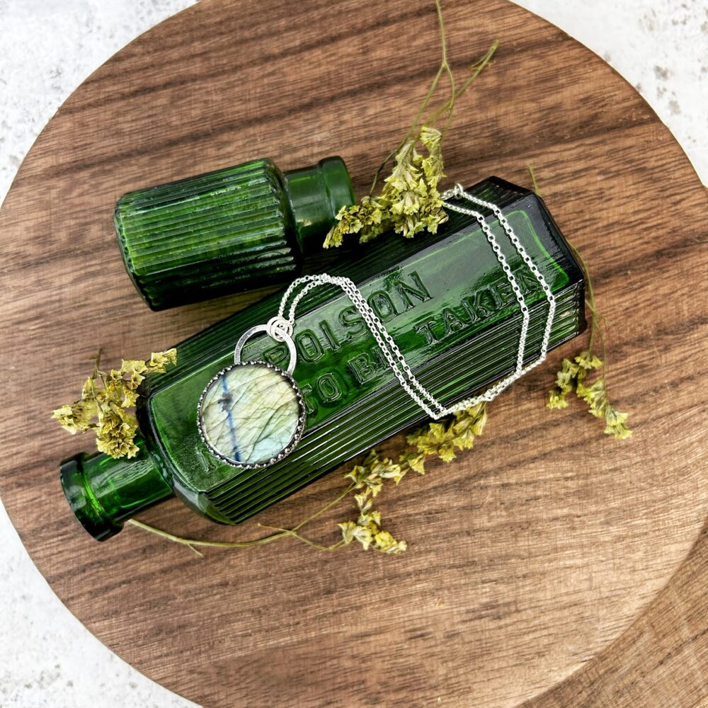 A large round labradorite necklace shown from above, displayed on a green Victorian bottle. The stone is a shimmering green colour and is set in a sterling silver bezel. Fran Barnett Jewellery