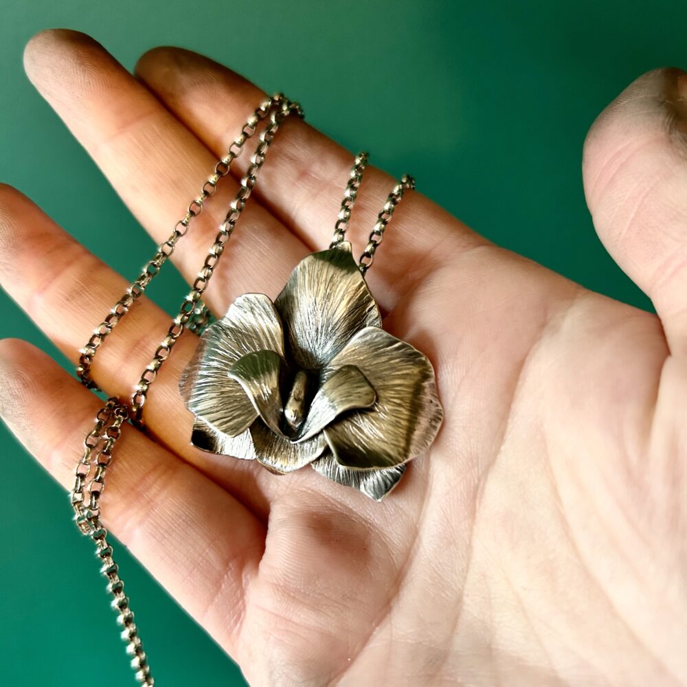 A large orchid pendant and chain, handmade from eco sterling silver. Held by a hand against a dark green background. Fran Barnett Jewellery