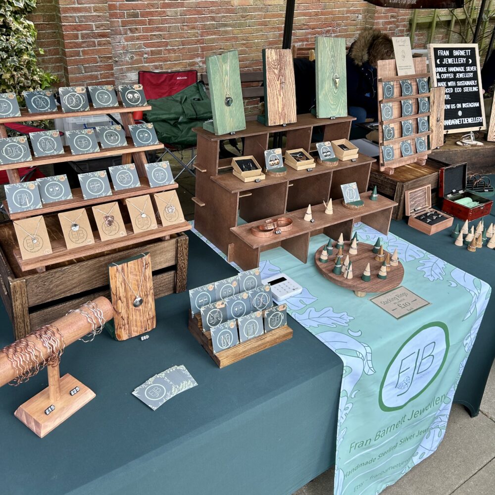 An outdoor market stall selling Fran Barnett Jewellery handmade sterling silver jewellery. The stall has dark green table cloths and a sage green table runner with the Fran Barnett Jewellery logo on it. The jewellery is displayed on rustic wooden stands