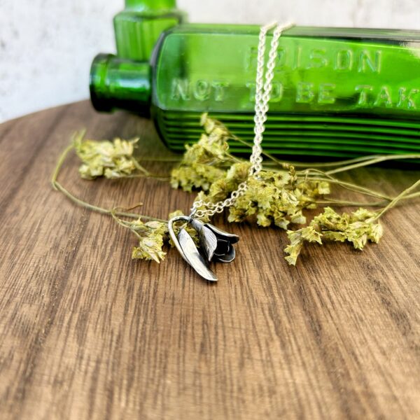 A delicate snowdrop handmade in eco silver hangs from a silver chain over an antique green glass bottle. Fran Barnett Jewellery