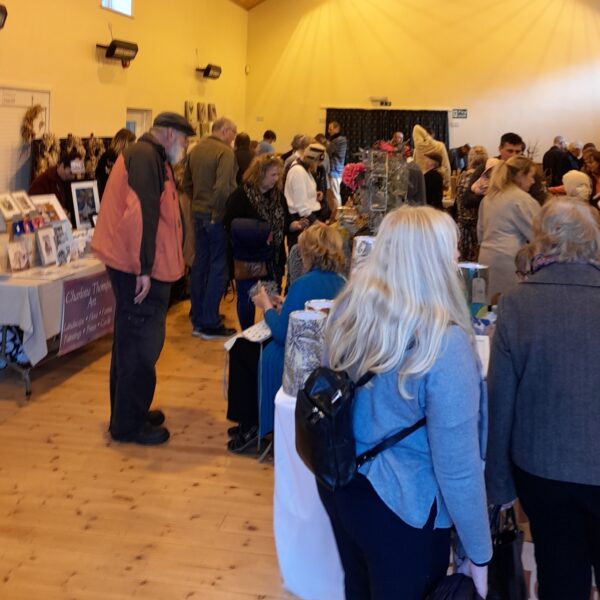 a photograph of a busy village hall, with customers looking at the different stall and speaking to makers.