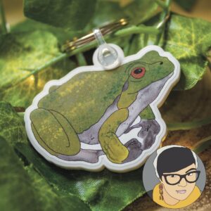 A recycled acrylic frog keychain sits on a next of leaves. In the bottom right corner there is my logo which is an illustration of me, a white woman with dark hair wearing a mustard yellow scarf and dark grey top.