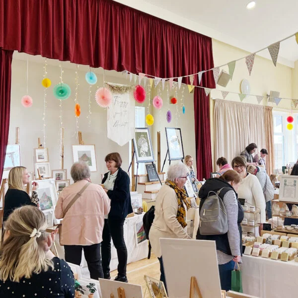 Customers are looking around a variety of market stalls in Worsall Village Hall. They are carrying shopping bags and chatting with stall holders. There are colourful decorations, bunting and fairy lights decorating the hall. There are lots of stalls selling handmade products including candles, artwork, jewellery, ceramics, glass, gifts, accessories, plants, and soap.