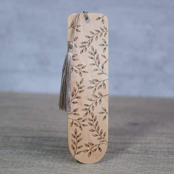 photo showing a wooden bookmark with a pyrography design. leaves cover the front of the bookmark. There is a dark green silky tassel attached to the top of the bookmark.