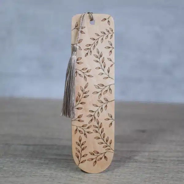 photo showing a wooden bookmark with a pyrography design. leaves cover the front of the bookmark. There is a dark green silky tassel attached to the top of the bookmark.