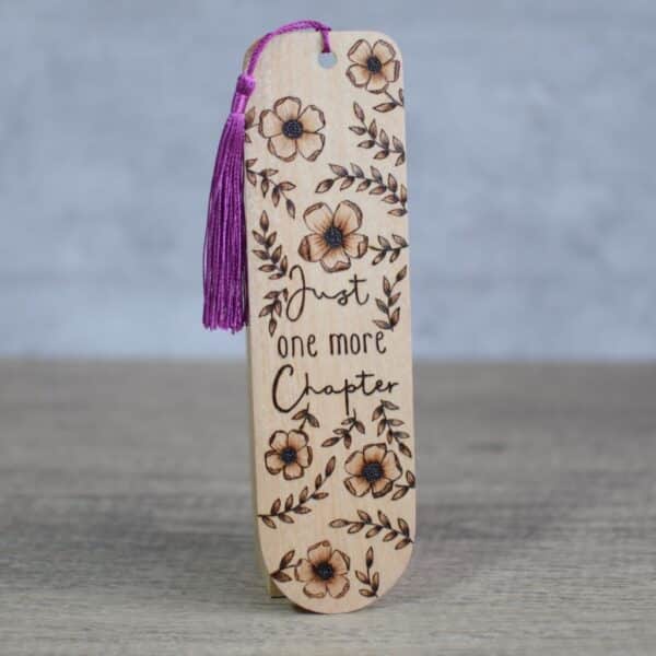 photo show a wooden bookmark with a floral and text design. the words "just one more chapter" are surrounded by blossoms and leaves.