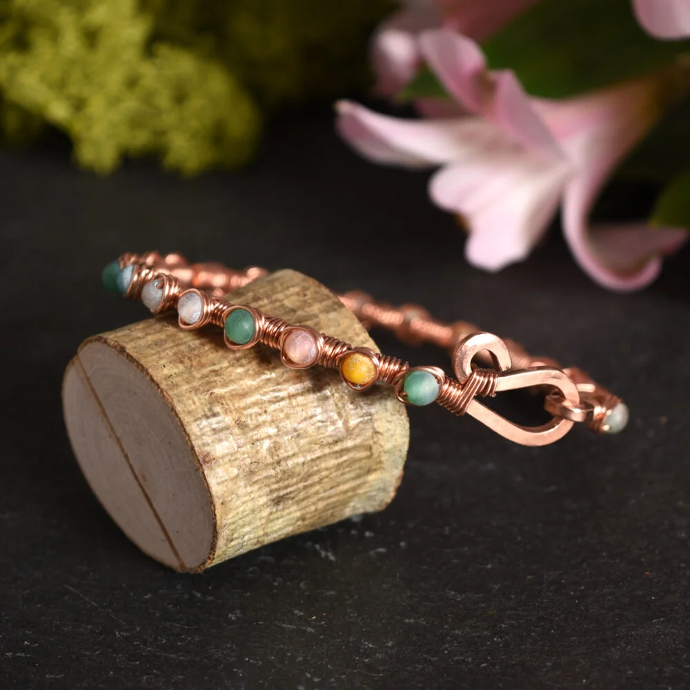 Wire Wrapped Multi Coloured Cracked Agate Copper Bangle propped on a small log against a black slate background with flower and moss in the background
