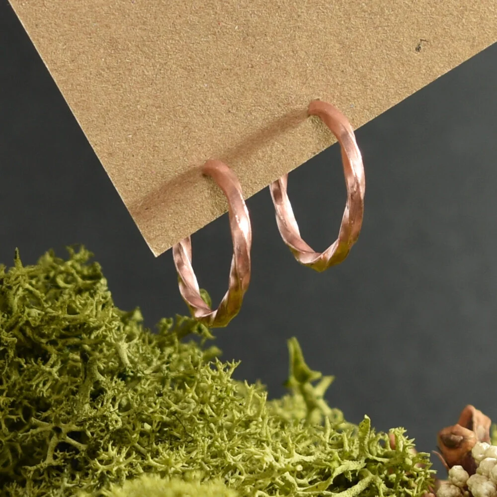 Twisted copper wire earrings suspended from a piece of natural card against a black background with green moss
