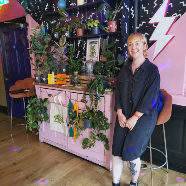 Chan from Sap Plants sat on a tall velvet stool next to a brigh pink and black retro 80's style dresser laiden with her plant stock and brightly coloured t-shit materiel pant hangers, tote bags and jewellery.