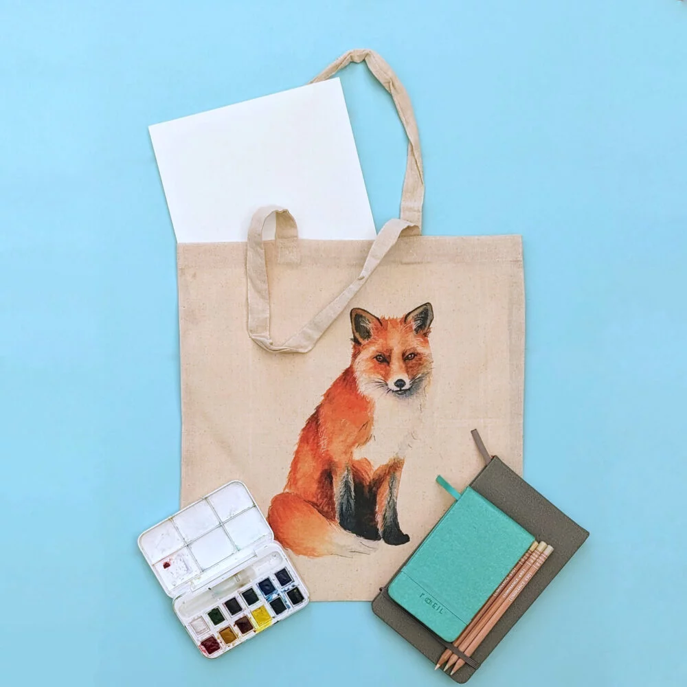 A tote bag with my Fox printed on with some art supplies surrounding it.