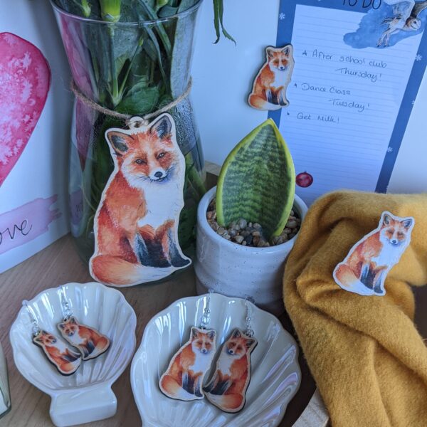 A hanging fox decoration on a vase of flowers, two sizes of fox earrings, a fox magent and a matching fox wooden pin badge
