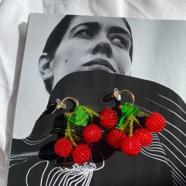 A pair of gold hoops attached to beaded red cherry earrings. Each hoop has four cherries, four stalks and two leaves. The earrings are placed on a clack and white magazine featuring the image of a woman. The magazine is on a white fabric background. Shop Gwynfor