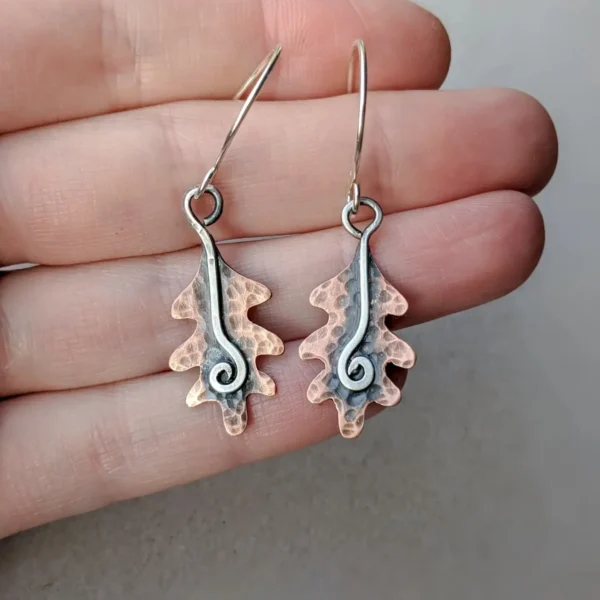 A hand holding a pair of copper earrings in the shape of an oakleaf, with silver wire decoration.
