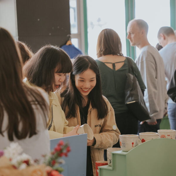 Two women are smiling together, looking at ceramics on a makers stall. The scene is at a market and behind them are people browsing other stalls, the room is bright.