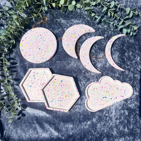A grey background with 4 white items with rainbow coloured chips in them. There is a cloud shaped tray, 2 hexagon shaped coasters and a wall hanging with 4 phases of the moon on it.