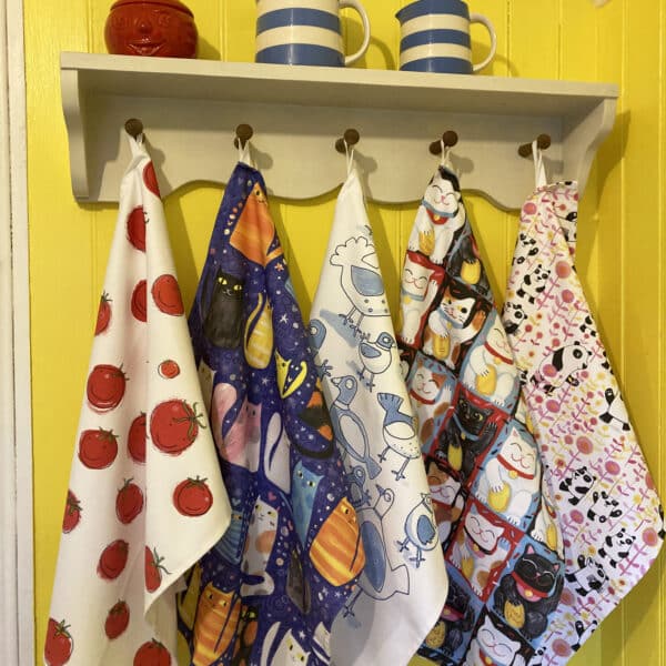 five different design printed tea towels hang from a peg rail with two blue striped jugs and a tomato shaped pot on top