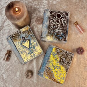 3 book shaped jewellery boxes in blue with moths, potion bottles and plants on the covers. Dotted around there are crystals and a large candle