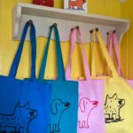 Four different coloured tote bags with dog illustrations hanging from a peg rail