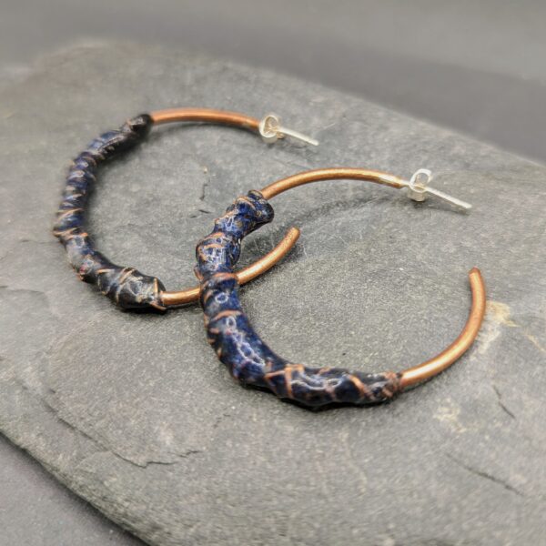 A pair of purple enamel and copper hoop earrings sit on a grey slate and background.