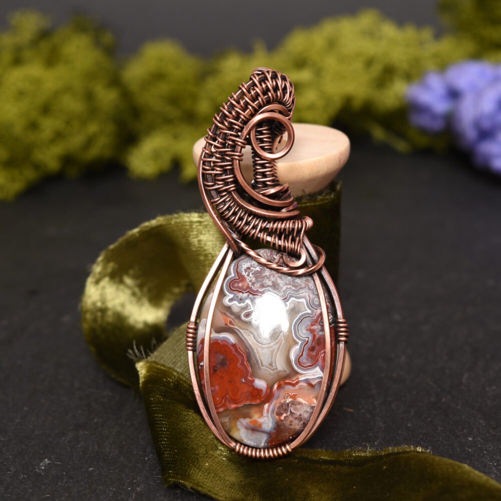 Lyn Haslegrave Jewellery - A red and cream crazy lace agate stone wire wrapped in antiqued copper wire displayed against an olive green ribbon spoil on a black background with natural green moss in the background