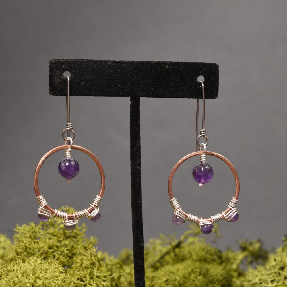 A pair of Copper hoop earrings featuring Amethyst gemstones and silver wire wrapping details hanging from a black display stand with natural green moss in the background
