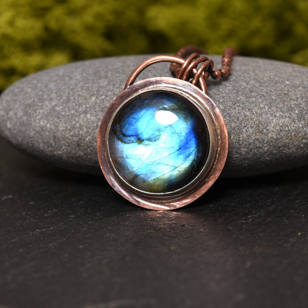 Round copper and silver labradorite pendant displayed propped against a natural grey stone. The background is black and features natural green moss
