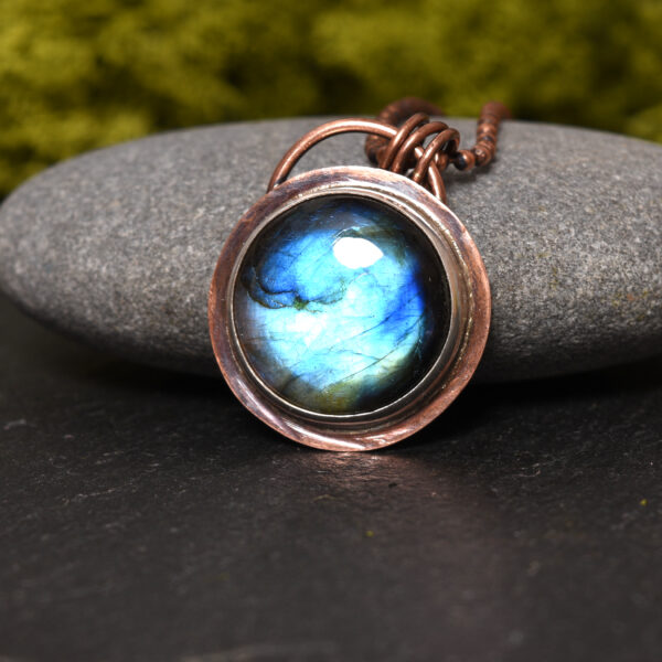 Round copper and silver labradorite pendant displayed propped against a natural grey stone. The background is black and features natural green moss
