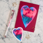 A Valentine card lays flat on a white marble coloured desk. The card features a red and blue watercolour painted heart on a deep pink background. A matching sticker lays on the desktop alongside it.