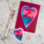 A Valentine card lays flat on a white marble coloured desk. The card features a red and blue watercolour painted heart on a deep pink background. A matching sticker lays on the desktop alongside it.