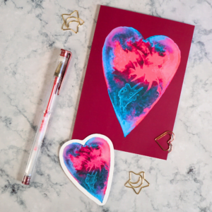 A Valentine card lays flat on a white marble coloured desk. The card features a red and blue watercolour painted heart on a deep pink background. A matching sticker lays on the desktop alongside it.