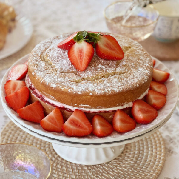 a home baked Victoria sponge cake sits on a white ceramic cake stand on top of a woven rattan mat. The cake has been dusted with white powdered sugar and decorated with sliced strawberries - there are three in a cluster on top of the cake and several more arranged in a ring around the base of the cake. Behind the cake in the background there is a glimpse of a pale, clear pink drink being poured into a clear, shimmery glass. There is also a glimpse of a pale, floral table cloth and some other baked goods on a plate.