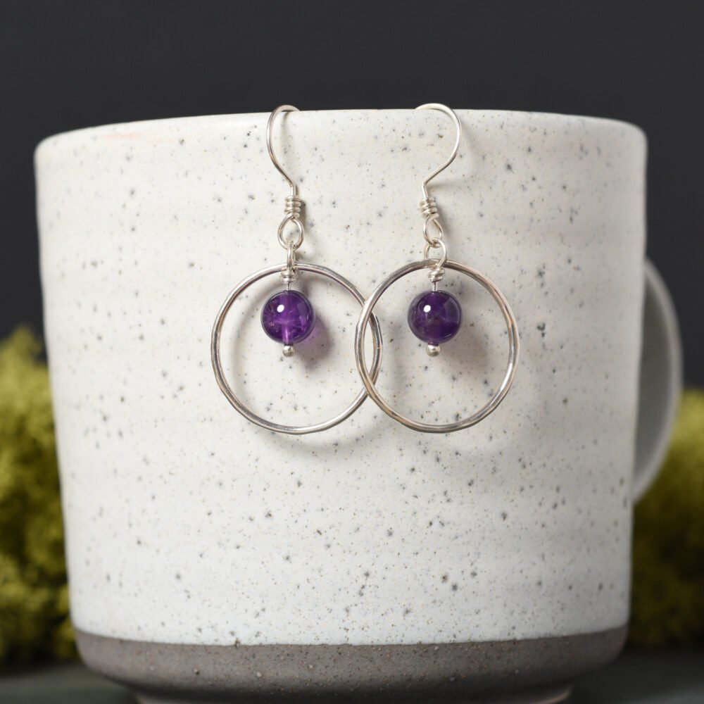 Silver hoops with an amethyst gemstone dangle in the middle suspended from silver hooks. Displayed hanging from a grey speckled mug with a black background featuring natural green moss
