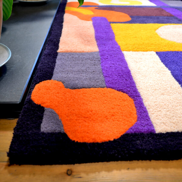 Image shows a large purple, orange, yellow, cream and burgundy handcrafted tufted wool rug, positioned in front of a log burner fireplace, with plants positioned at the sides. Rug is placed on wooden floorboards.