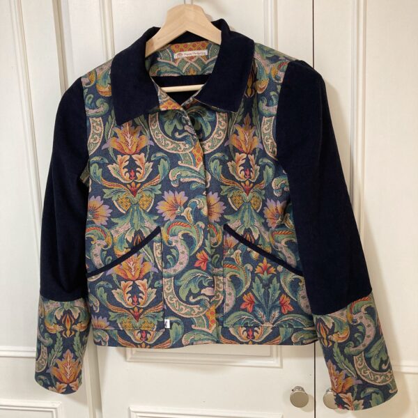 A woman's jacket hanging against a white cupboard. The jacket has a colourful vintage botanical print on the front of the jacket & contrast sleeves and collar in navy corduroy. Handmade by Happy Hedgehog Designs