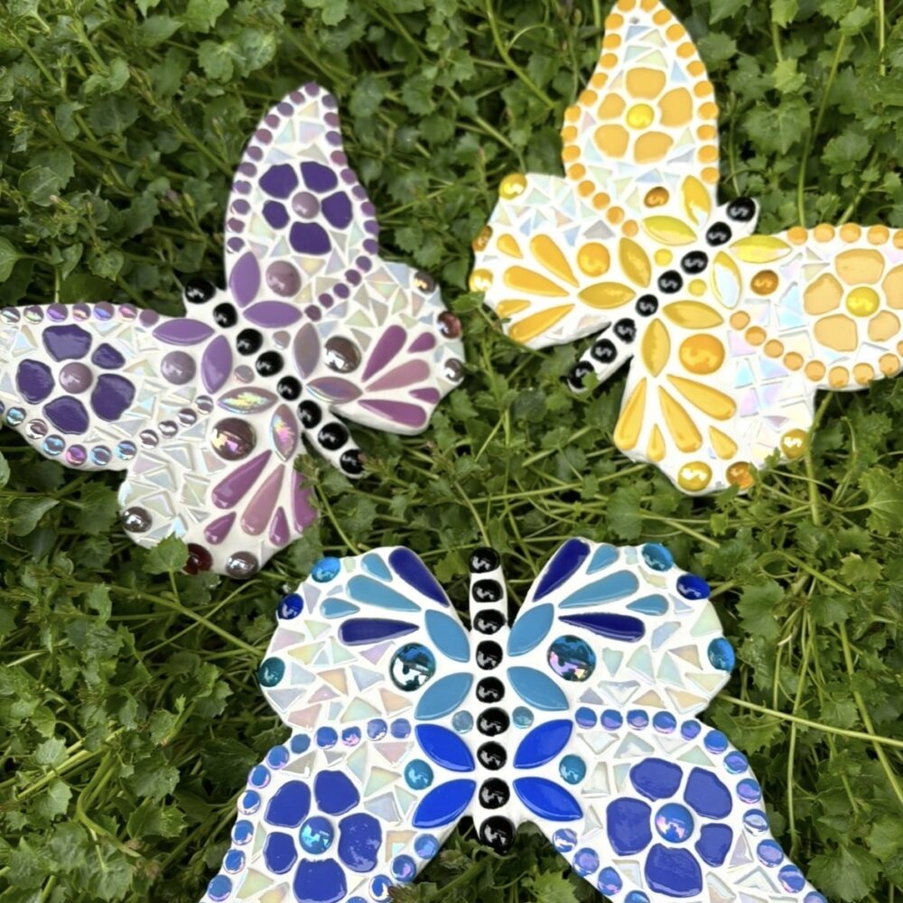Three handmade mosaic butterflies resting on lush green foliage. The butterflies feature purple, yellow and blue glass mosaic designs with iridescent tiles, glossy black bodies and intricate decorative patterns. Colourful and nature-inspired, the butterflies showcase detailed craftsmanship and vibrant handmade mosaic art by Debbee at Mom’s Mosaics