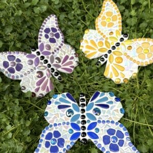 Three handmade mosaic butterflies resting on lush green foliage. The butterflies feature purple, yellow and blue glass mosaic designs with iridescent tiles, glossy black bodies and intricate decorative patterns. Colourful and nature-inspired, the butterflies showcase detailed craftsmanship and vibrant handmade mosaic art by Debbee at Mom’s Mosaics