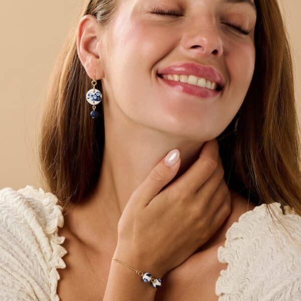 a female model wearing a pair of blue and white floral dangle earrings and a dainty floral bracelet