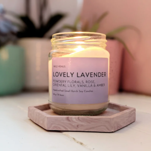 Lovely lavender candle by Wild Venus. The Lavender and vanilla scented candle is sitting on a pink coaster with plants in the background.