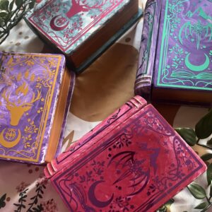 A close up of 4 book shaped jewellery boxes with shiny colours of vinyl on them. They are pink, purple, teal and gold